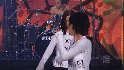 Man Playing Drums Behind Two Women, One Wearing Hate Shirt GIF