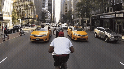 Man Riding Bike Down Street With Taxis And Bank Of America GIF