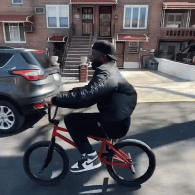 Man Riding Red Bike Down Street Past Brick Building GIF