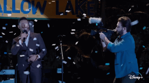 Man Singing Into Microphone In Front Of Allow Lake Sign GIF