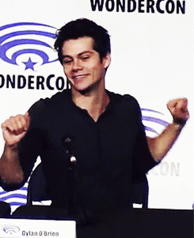Man Sitting At Table With Arms In Air In Front Of Wondercon Sign GIF