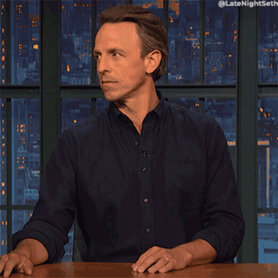 Man Sitting At Table With Hands Outstretched In Front Of Late Night Seth Screen GIF