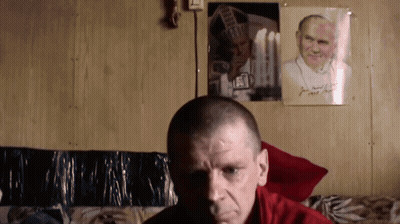 Man Sitting In Front Of Wall With Picture And John Paul Sign GIF