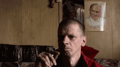 Man Smoking Cigarette In Front Of Picture Named John GIF