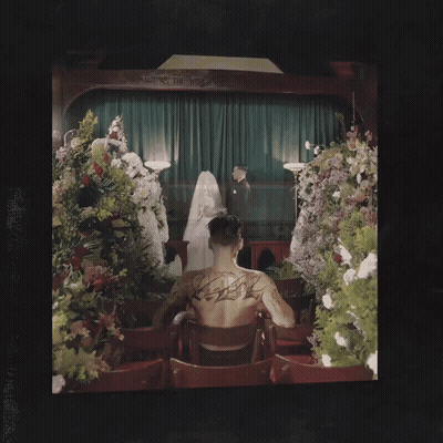 Man With Back Tattoo Beside Coffin And Flowers GIF