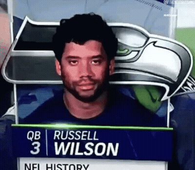 Man With Beard Standing In Front Of Russell Wilson Sign GIF