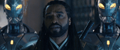 Man With Dreadlocks Standing Before Two Robots The Illuminati Above GIF