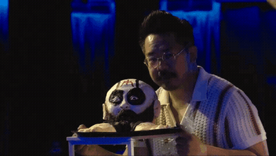 Man With Glasses Holding Puppet With Cross On Face GIF