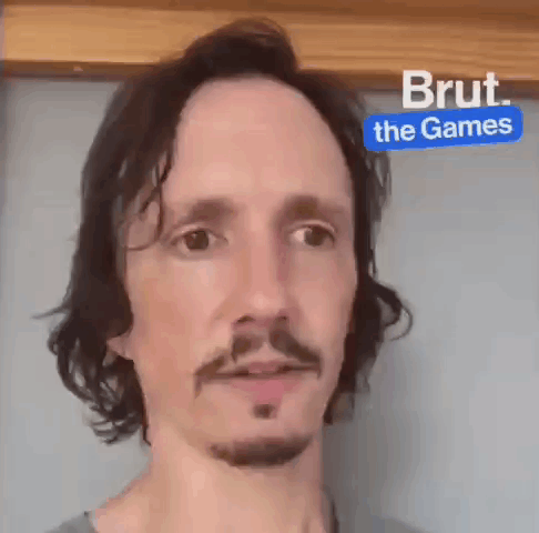 Man With Long Hair And Beard In Front Of Blue Sign Saying Brut The Games GIF