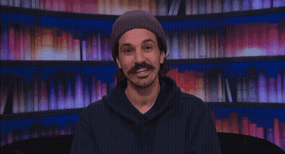 Man With Mustache In Beanie And Hoodie Smiling By Bookshelf GIF