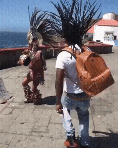 Man With Orange Mcm Backpack Next To Man In Native American Outfit GIF