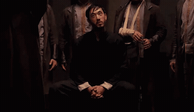 Men Standing Around Seated Man In Dark GIF