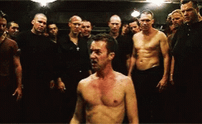 Men Standing Around Shirtless Man In Dark Room GIF