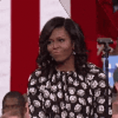 Michelle Obama In Black And White Polka Dot Dress In Front Of Red And White Flag GIF