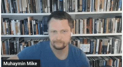 Muhaymin Mike Is Shown In Front Of A Library Full Of Books GIF