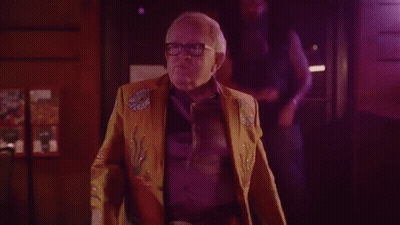 Older Man In Yellow Jacket And Glasses In Dark Room GIF