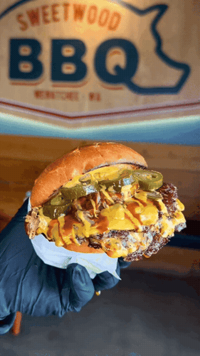 Person Holding Hamburger In Front Of Sweetwood BBQ Sign GIF