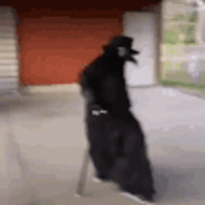 Person In Black Cloak And Hat Walking Down Sidewalk GIF