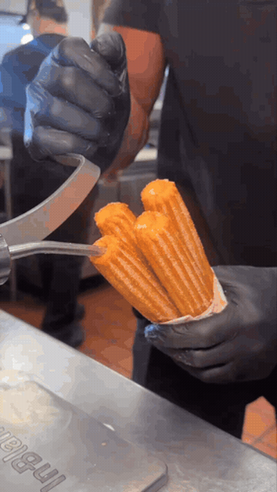 Person In Black Gloves Holding Churro In Front Of I-Ball Sign GIF