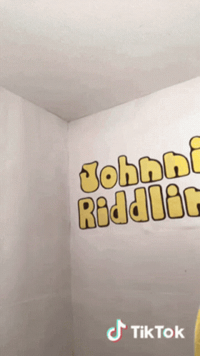 Person In Yellow Shirt In Front Of Johnny Riddler Wall GIF