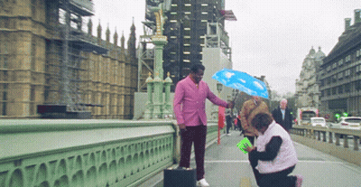 Pink-Suited Man Holds Blue Umbrella Over Woman Bridge GIF