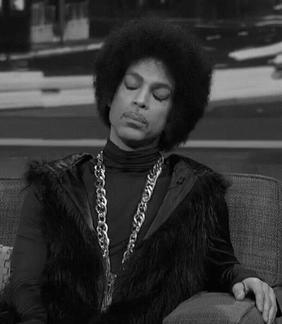Prince Is Sitting On A Couch Wearing A Fur Vest And A Chain . GIF