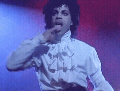 Prince Is Wearing A White Shirt And Ruffled Collar And Is Sticking His Tongue Out While Dancing On Stage . GIF