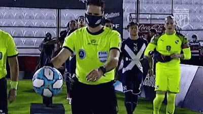 Referee With Face Mask Holding Soccer Ball In Front Of Players GIF