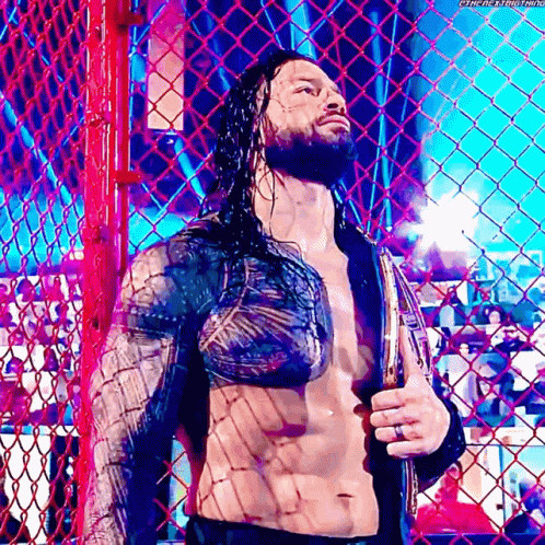 Roman Reigns Is Standing Behind A Chain Link Fence Holding A Belt . GIF