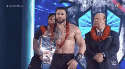 Roman Reigns Is Wearing A Lei And Holding A Wrestling Championship Belt While Standing On A Stage . GIF