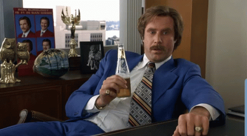 Ron Burgundy Is Sitting At A Desk With A Bottle Of Beer In His Hand GIF