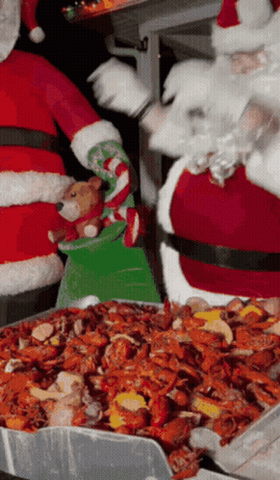 Santa Claus Is Holding A Stuffed Animal While Standing Next To A Tray Of Crawfish GIF