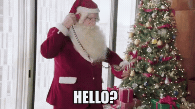 Santa Claus Is Talking On A Phone In Front Of A Christmas Tree And Says Hello ? GIF