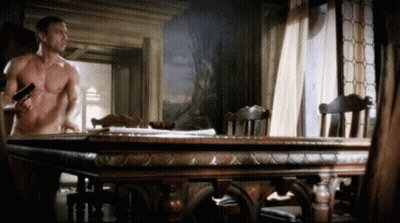 Shirtless Man Holding Gun In Front Of Wooden Table GIF
