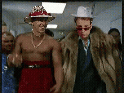 Shirtless Man In Cowboy Hat And Fur Coat Next To Another Man GIF