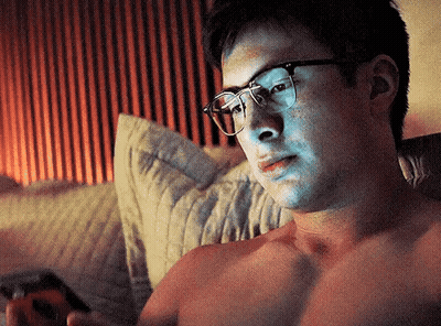 Shirtless Man With Glasses Laying On Bed Looking At Phone GIF