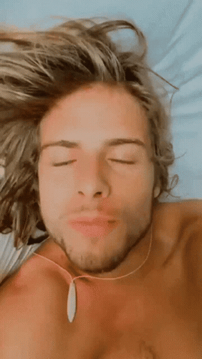 Shirtless Man With Long Blonde Hair Laying On Bed Eyes Closed Necklace GIF