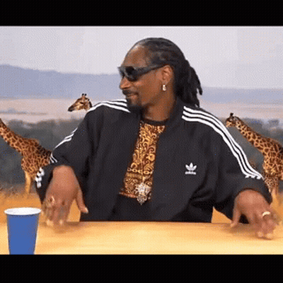 Snoop Dogg Is Sitting At A Table With A Blue Cup And Giraffes In The Background . GIF