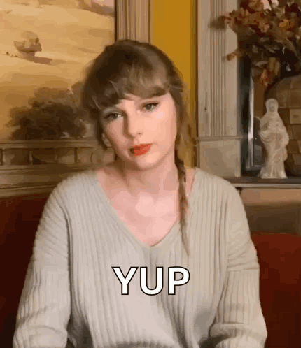 Taylor Swift Is Wearing A Grey Sweater And Sitting On A Red Couch With A Statue In The Background . GIF