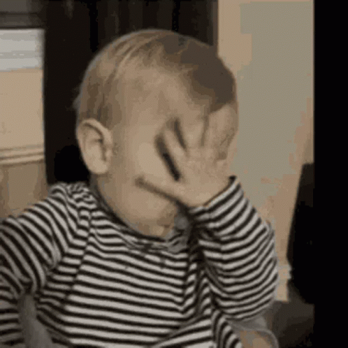 The Baby Is Wearing A Striped Shirt And Covering His Face With His Hand . GIF