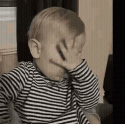 The Baby Is Wearing A Striped Shirt And Covering His Face With His Hand . GIF
