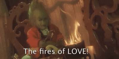 The Grinch Is Sitting In Front Of A Fireplace With The Words `` The Fires Of Love '' Written On The Screen . GIF