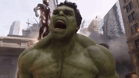 The Hulk Is Standing In The Middle Of A City With His Mouth Open And A Statue Of Iron Man In The Background . GIF