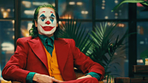 The Joker Is Sitting In A Chair With His Hands Folded GIF