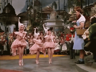 Three Ballerinas In Pink Tutus Are Dancing In Front Of A Crowd While A Woman In A Blue Dress Stands Behind Them . GIF