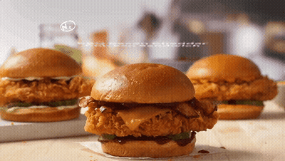 Three Fried Chicken Sandwiches On A Wooden Table With A N On The Bottom Right GIF