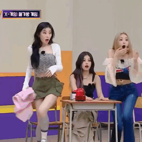 Three Girls Are Sitting At Desks In A Room With A Sign That Says X-Game On It . GIF