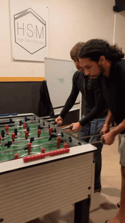 Three Men Are Playing A Game Of Foosball In Front Of A Sign That Says Hsm GIF