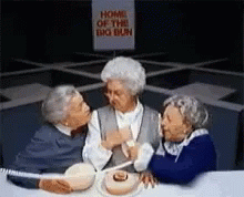 Three Older Women Are Sitting At A Table With A Sign That Says Home Of The Big Bun GIF
