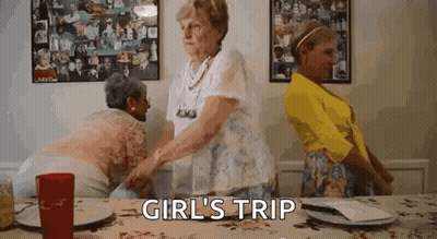 Three Older Women Are Standing Around A Table With The Words `` Girl 'S Trip '' Above Them . GIF